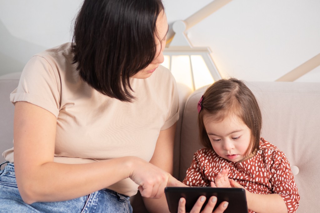 A woman and a young girl sitting on a couch together, looking at and interacting with a tablet